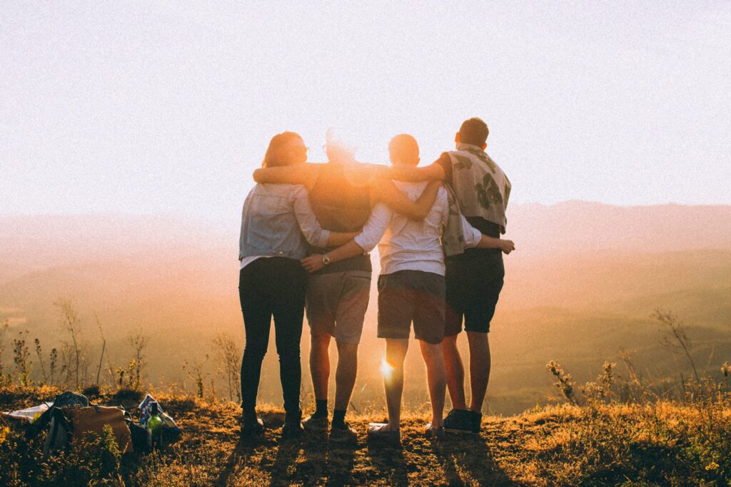 Four friends with arms around each other look out over a wide valley as the sun glows in the distance