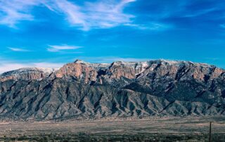 A ridge of mountains, the Sadia Crest in Albuquerque, New Mexico, with a deep blue sky and high, light clouds overhead and the dry valley floor in the foreground