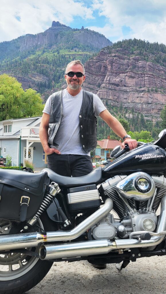 Joe LaMastra standing behind a Harley Davidson motorcycle in Ouray, Colorado, with pine covered rocky mountains in the background and bright blue, partly cloudy sky.